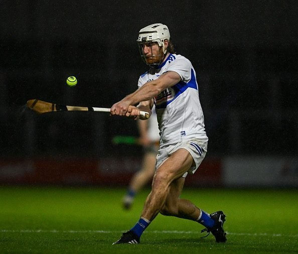 A hurler strikes an Illuminous sliotar under floodlights on a wet night